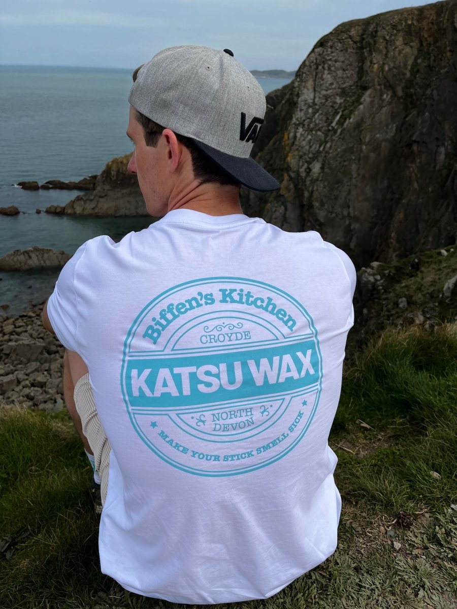 Person wearing a white t-shirt with 'Katsu Wax' branding by the sea.