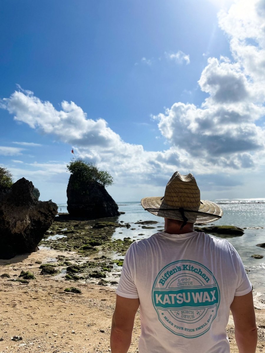 Person wearing a straw hat and Katsuwax t-shirt on a beach with rocks and ocean in the background