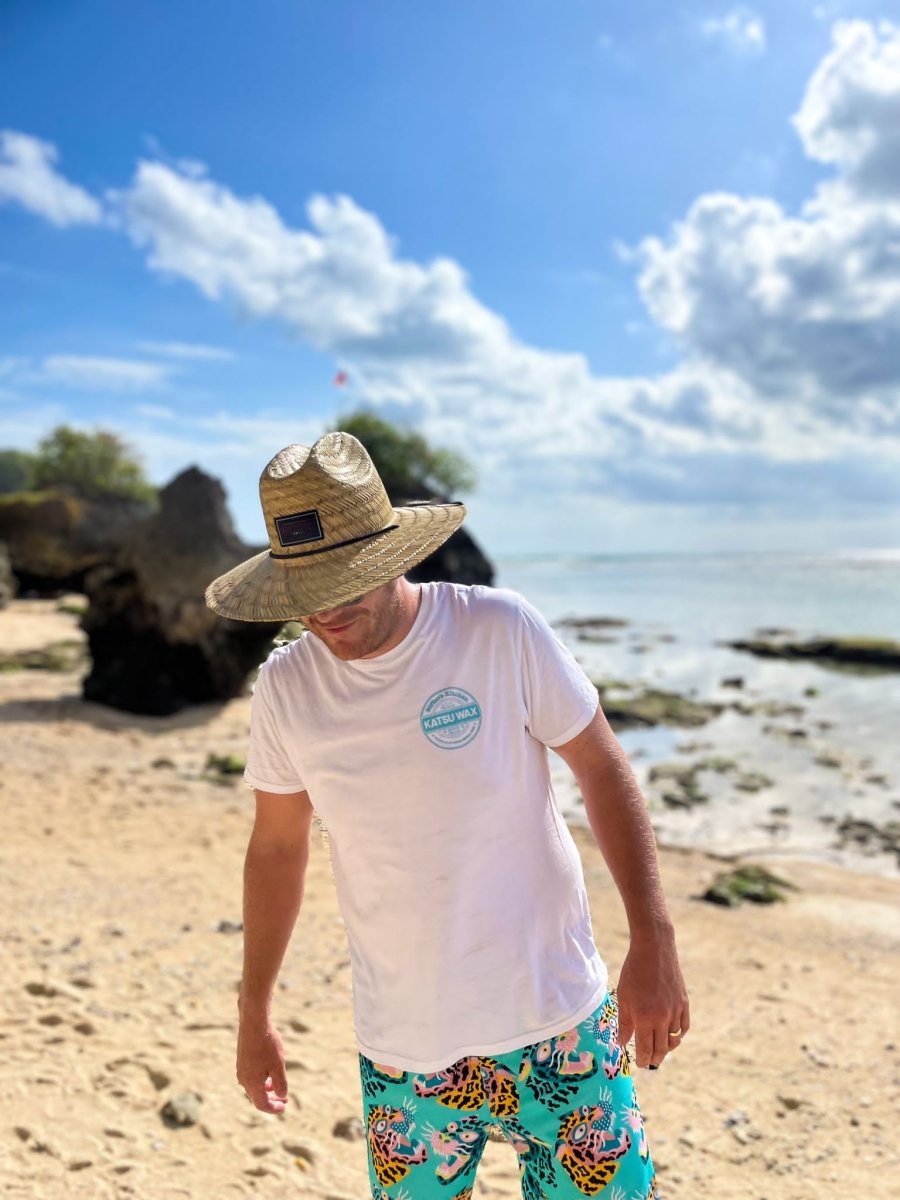 Man wearing a straw hat, white t-shirt, and colorful shorts on a beach.