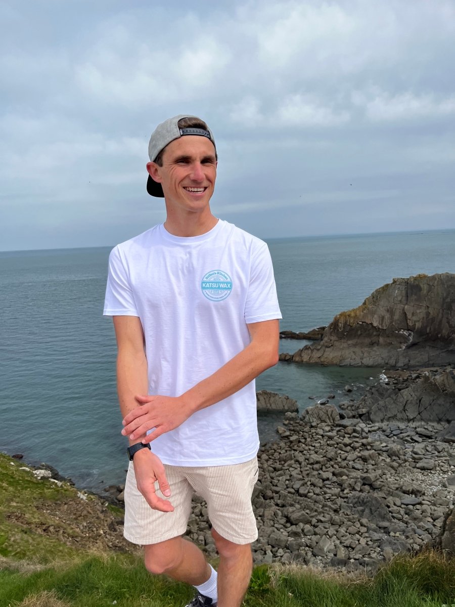 Man standing on a rocky shore with ocean view wearing a white t-shirt and cap.